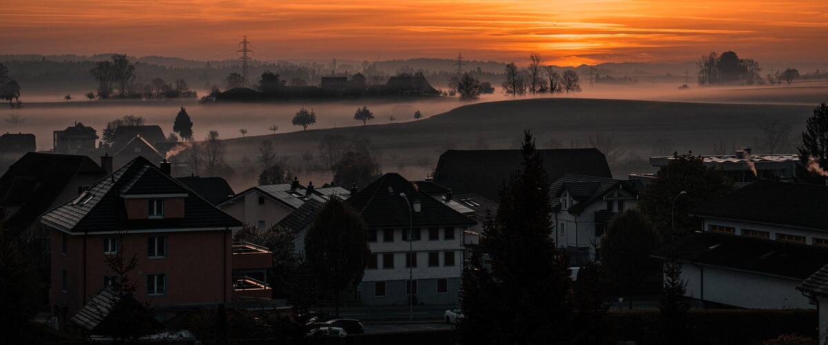 A sunrise over a misty landscape with houses and a power line tower.