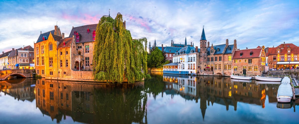 Uma cidade histórica europeia com um rio calmo, refletindo edifícios e uma imponente árvore de salgueiro-chorão.