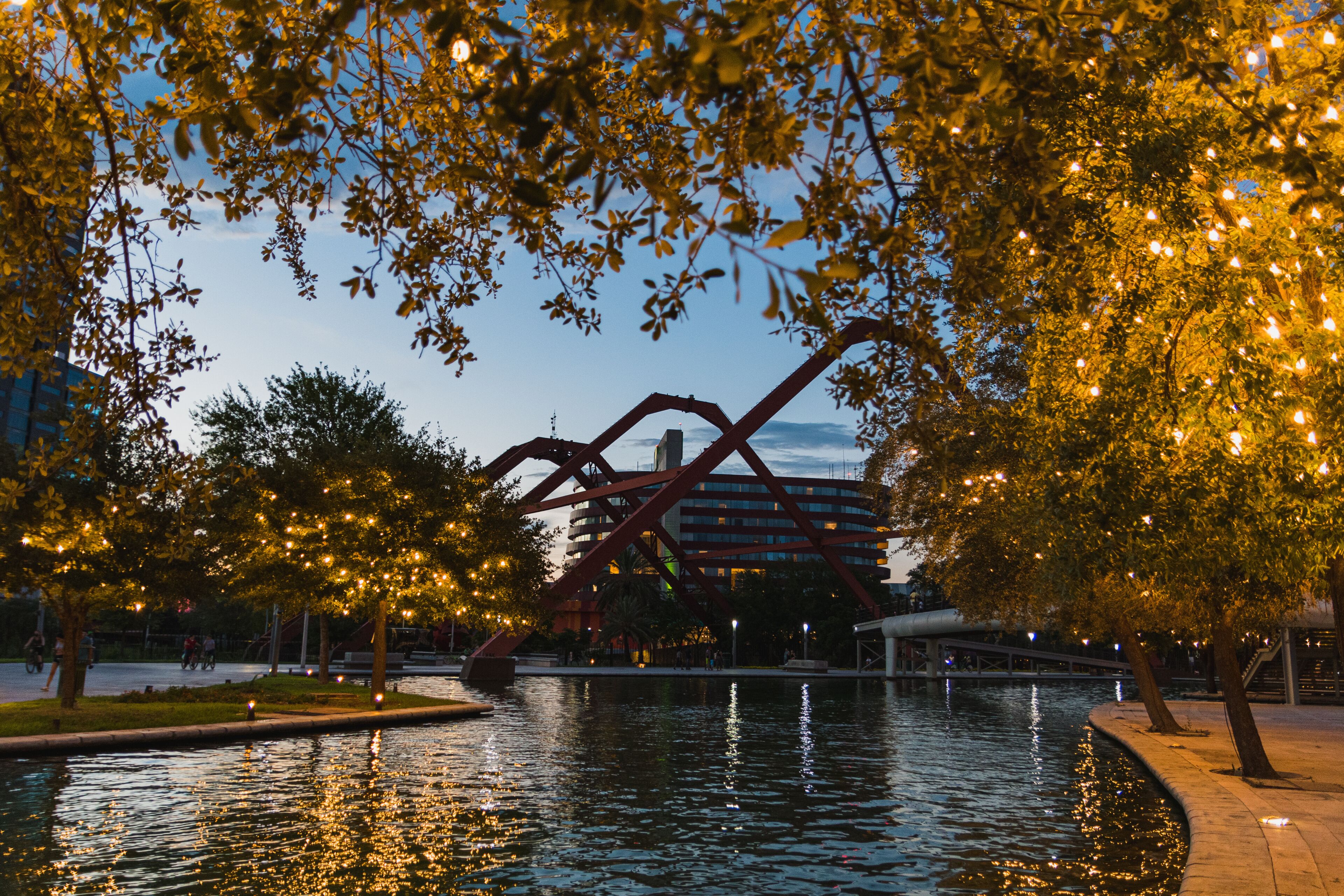 Um parque com um corpo d'água reflexivo, árvores iluminadas e um edifício moderno ao fundo.