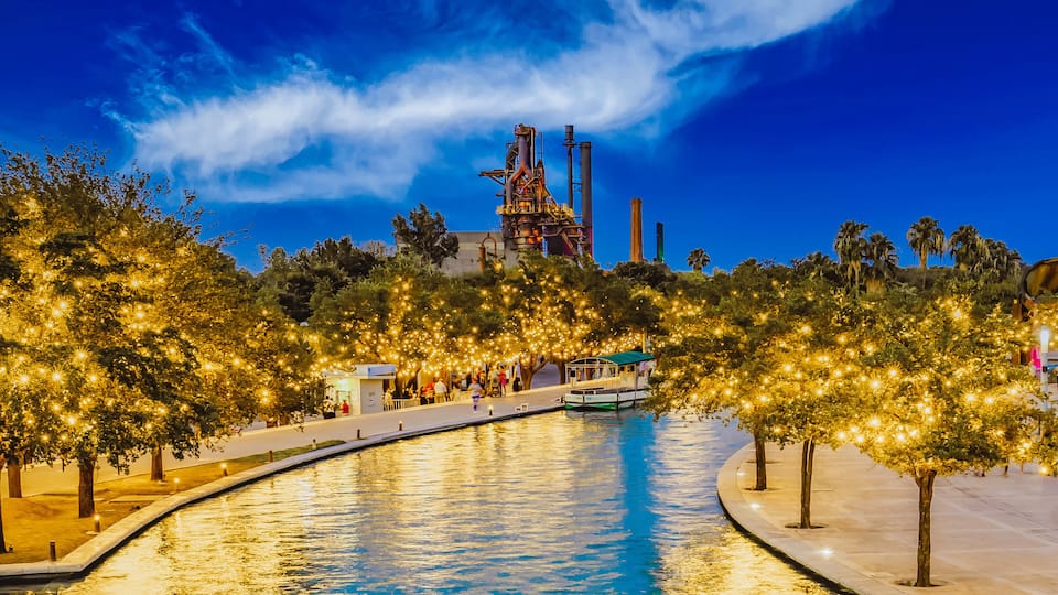 A nighttime riverside scene with illuminated trees, a calm water body, and industrial structures in the background.