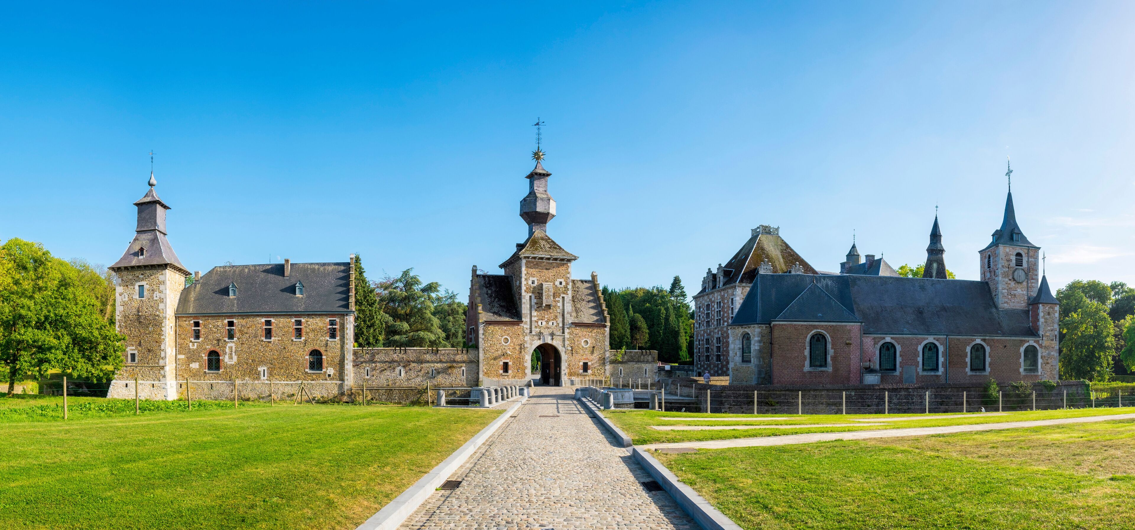 Um castelo histórico com várias torres e um portão central, rodeado por um jardim bem cuidado e caminhos de paralelepípedos.