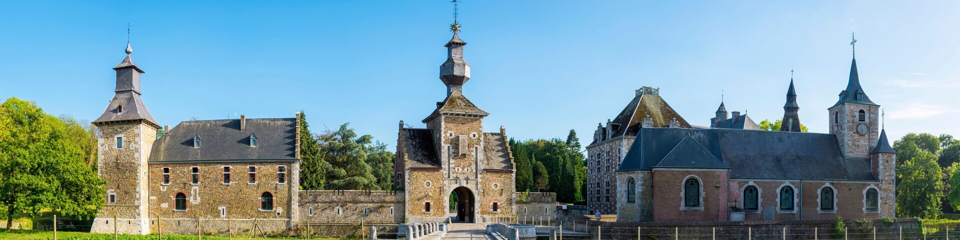 Um castelo histórico com várias torres e um portão central, rodeado por um jardim bem cuidado e caminhos de paralelepípedos.