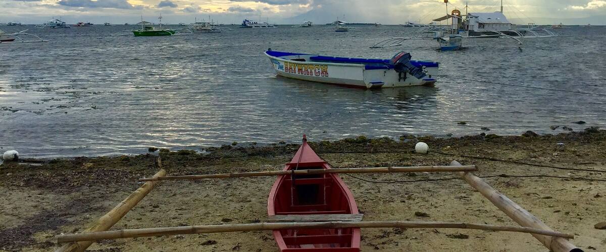A beach with a small red boat in the foreground and a larger boat named "The Bay Watch" in the background.