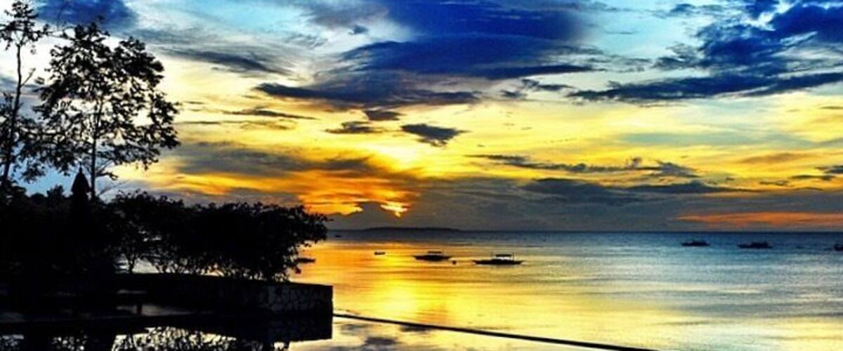 A sunset over a calm sea with boats, a reflective water surface, and silhouettes of trees.