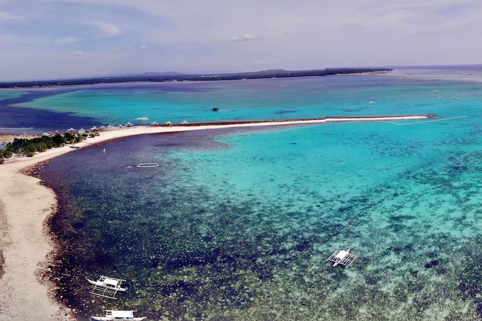 Vista aerea di una zona costiera con acque turchesi cristalline, una spiaggia sabbiosa e una piccola isola con vegetazione.