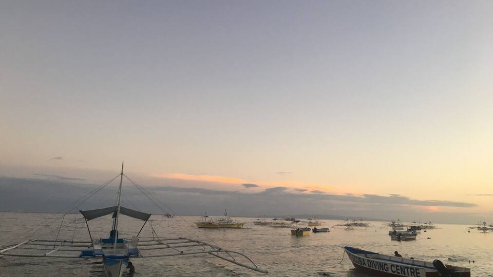 Une mer calme où l’on aperçoit plusieurs bateaux, dont un portant l’inscription « CENTRE DE PLONGÉE SOUS-MARINE », ainsi qu’un bateau traditionnel à voile.