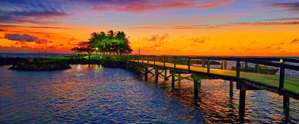 Un muelle de madera que se extiende sobre aguas tranquilas, palmeras y un cielo al atardecer.