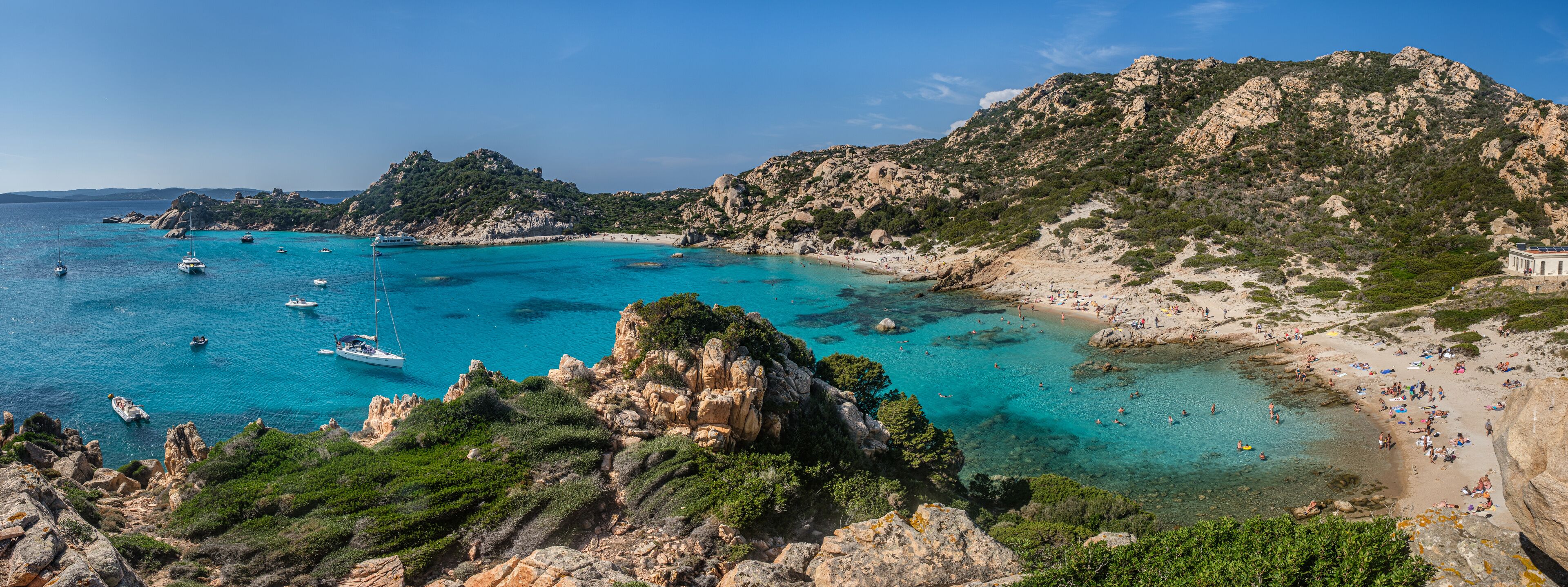 A coastal landscape with clear turquoise waters, rocky cliffs, and a sandy beach populated with people.
