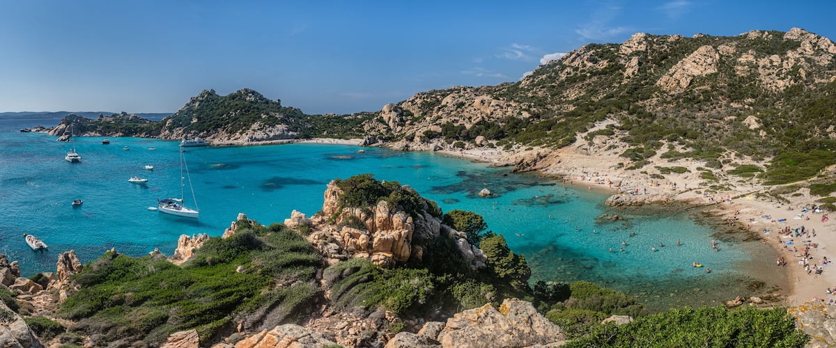 A coastal landscape with clear turquoise waters, rocky cliffs, and a sandy beach populated with people.