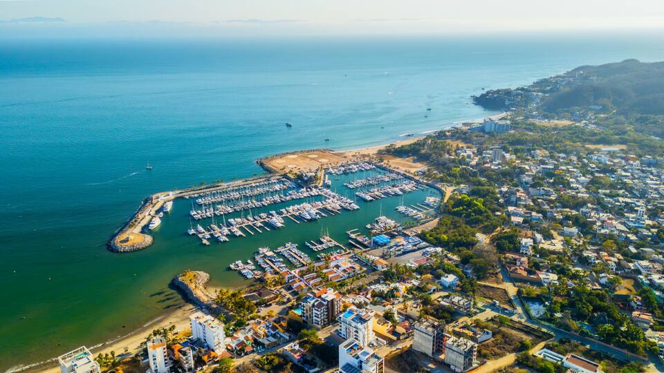 An aerial view of a coastal marina with numerous boats docked, surrounded by residential buildings and a hilly landscape in the background.