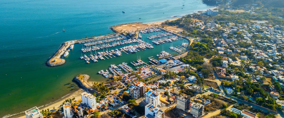 An aerial view of a coastal marina with numerous boats docked, surrounded by residential buildings and a hilly landscape in the background.