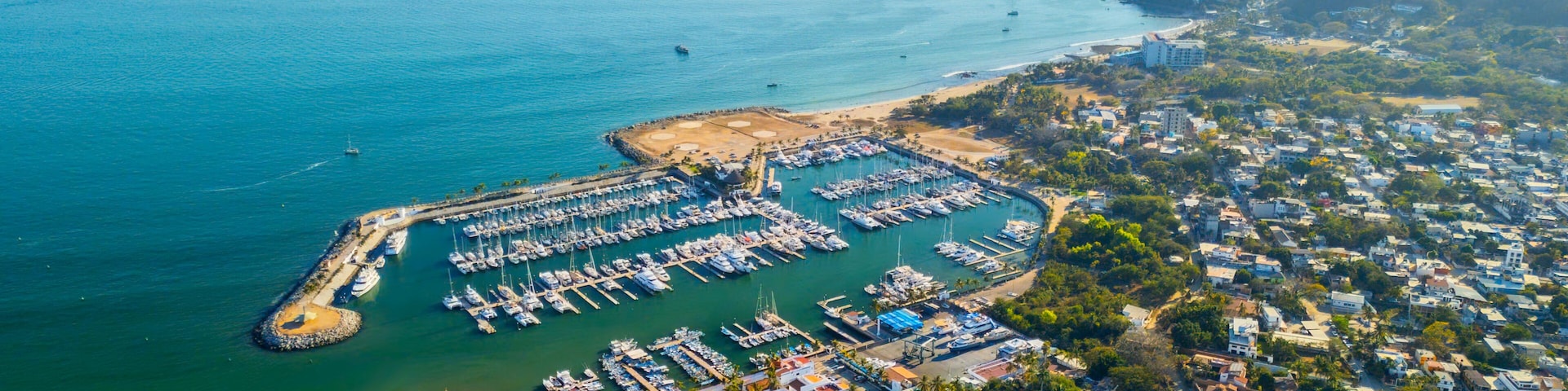 An aerial view of a coastal marina with numerous boats docked, surrounded by residential buildings and a hilly landscape in the background.