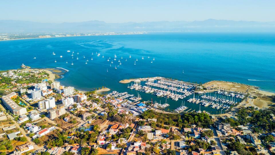 A coastal marina with numerous boats, a densely built urban area, and a clear view of the surrounding mountains.