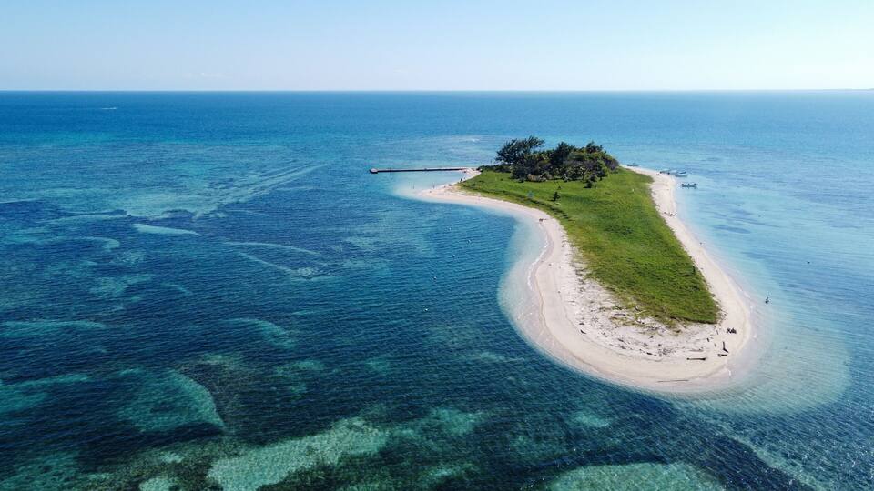 An aerial view of a narrow island with a beach, surrounded by clear blue waters.