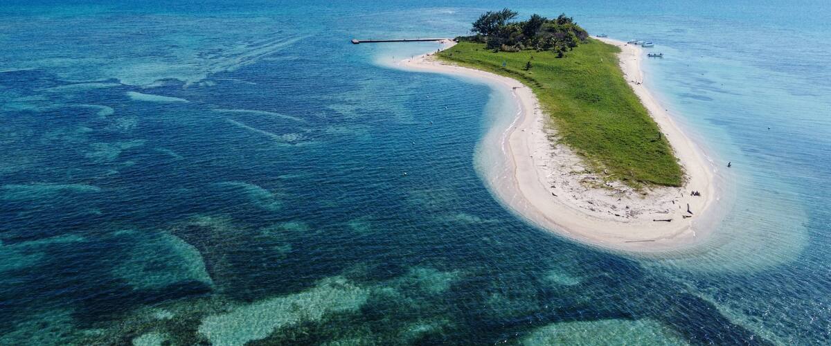An aerial view of a narrow island with a beach, surrounded by clear blue waters.