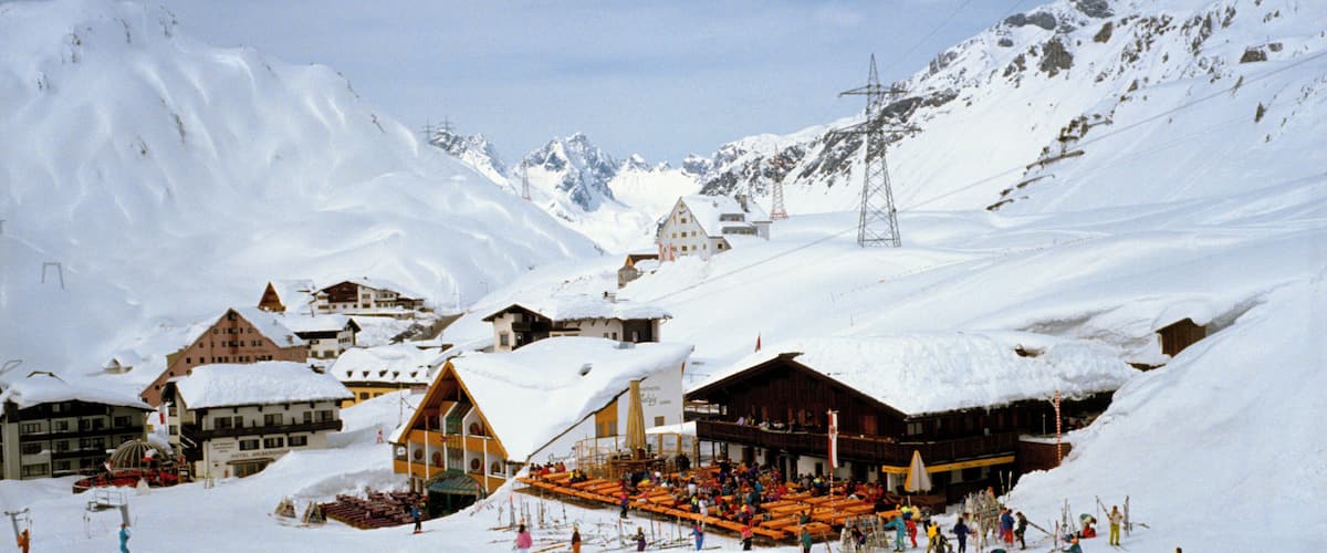 A snow-covered ski resort with buildings, a ski lift, and a mountainous backdrop.