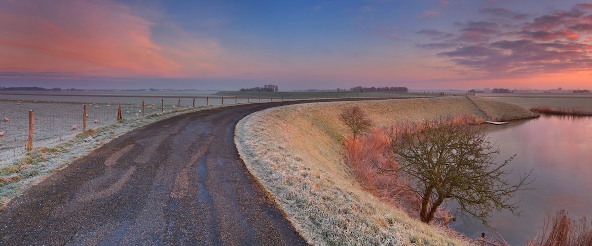 A rural landscape with a road curving alongside a body of water, fields, and a fence with sheep.