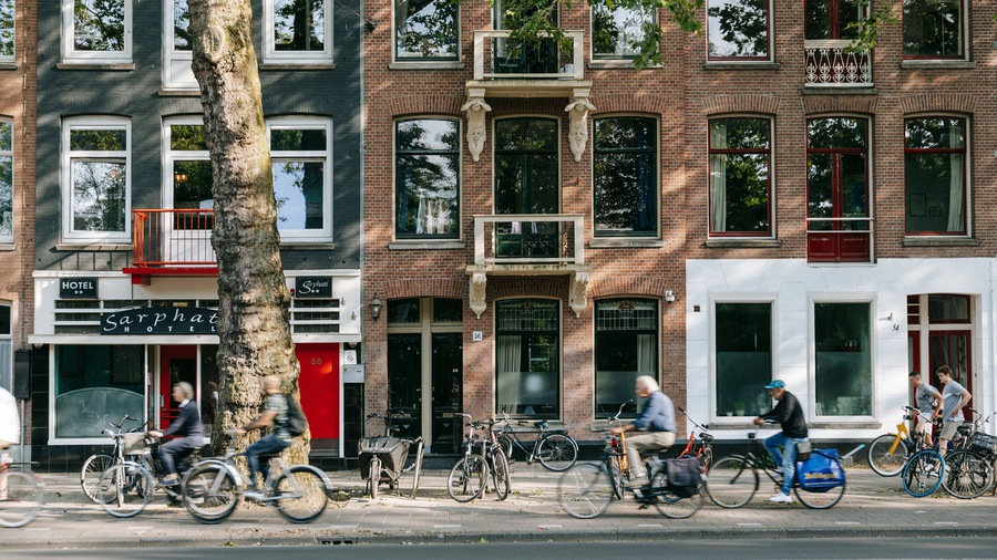 Een straat met fietsen langs de stoep, een boom en gebouwen met borden voor een hotel en een restaurant.