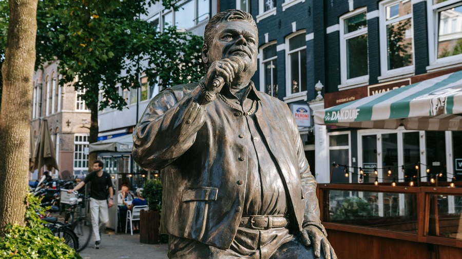Een bronzen beeld van een man die een microfoon vasthoudt, met een boom en gebouwen op de achtergrond.