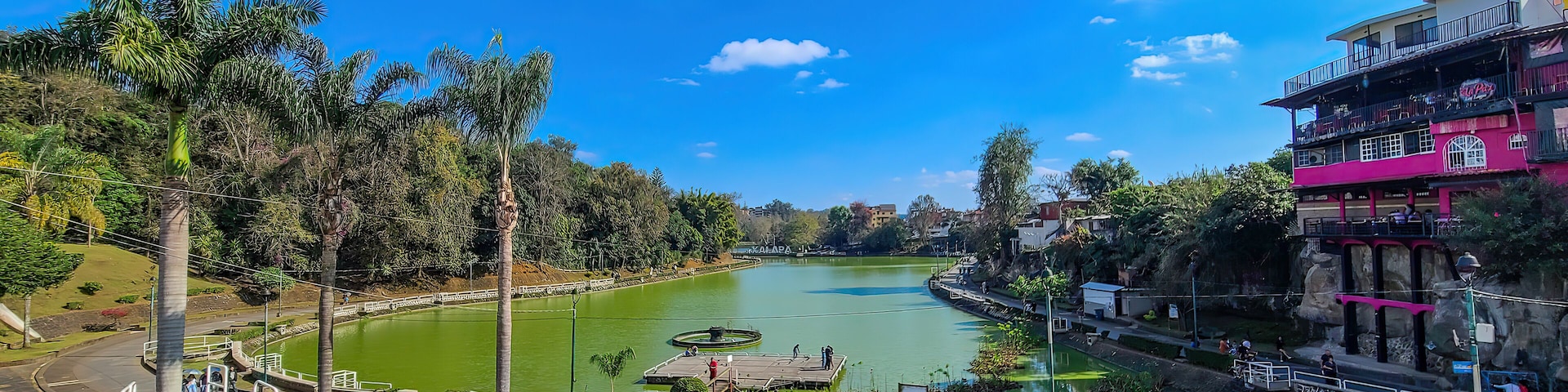 A lake with a dock, surrounded by trees and a pink building.