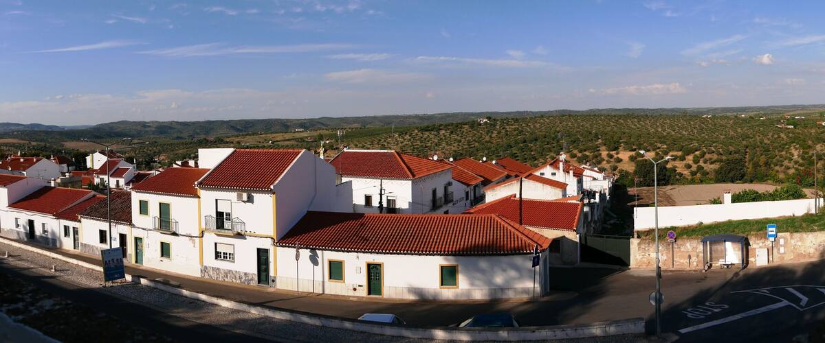 A coastal town with white buildings and red roofs, a clear sky, and a hilly landscape in the background.