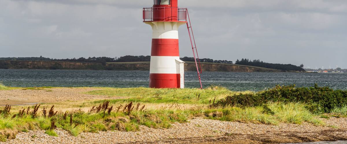 A red and white lighthouse on a rocky shore with a view of the sea and distant land.