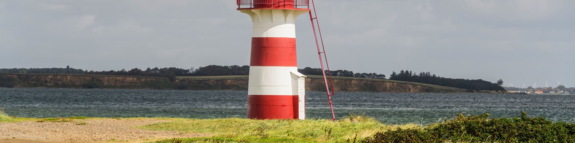 A red and white lighthouse on a rocky shore with a view of the sea and distant land.