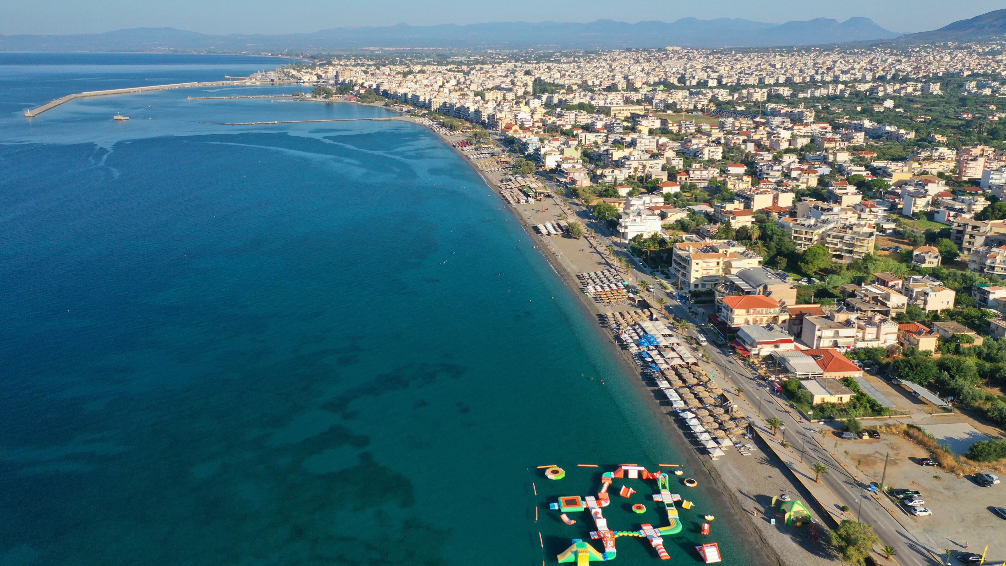 An aerial view of a coastal city with a long beachfront, dense urban area, and a pier with colorful play structures.