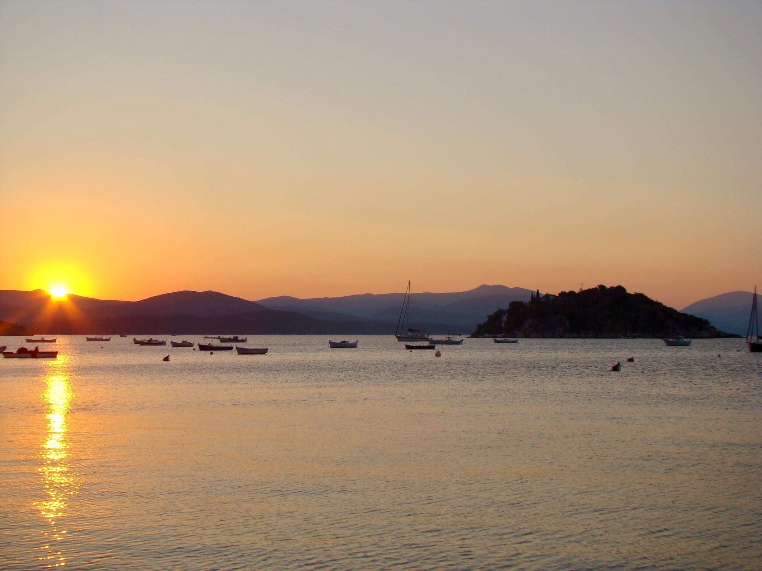 A sunset over a calm sea with several boats and a distant hill.
