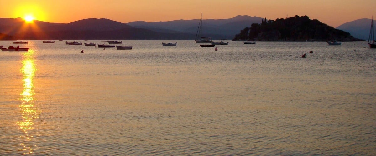 Un coucher de soleil sur une mer calme, avec plusieurs bateaux et une colline au loin.