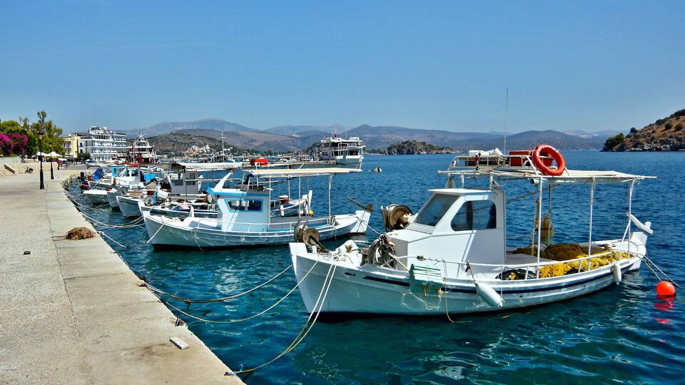 A marina with several small boats docked along a concrete quay, with buildings and hills in the background.