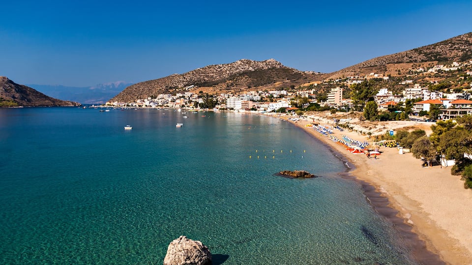 A coastal town with clear blue waters, a sandy beach, and buildings along the shore.