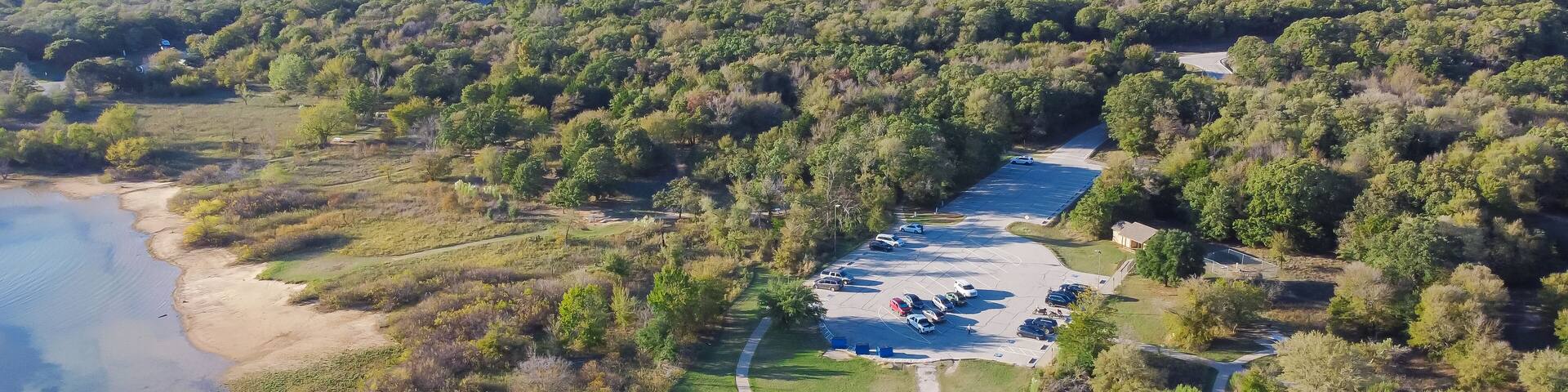Park entrance parking lots with parked cars, restroom in lush green tree forest of Isle du Bois Ray Roberts Lake State Park, water tower Denton in background, camping sites