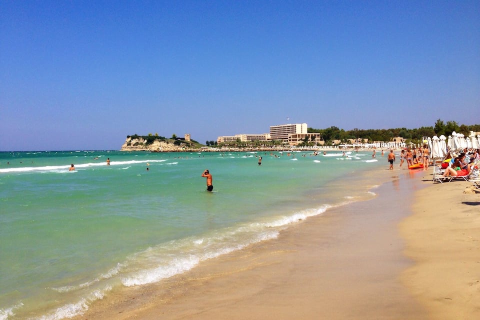Une plage à l’eau turquoise et limpide, une rive sablonneuse et, au loin, une construction qui ressemble à un château.