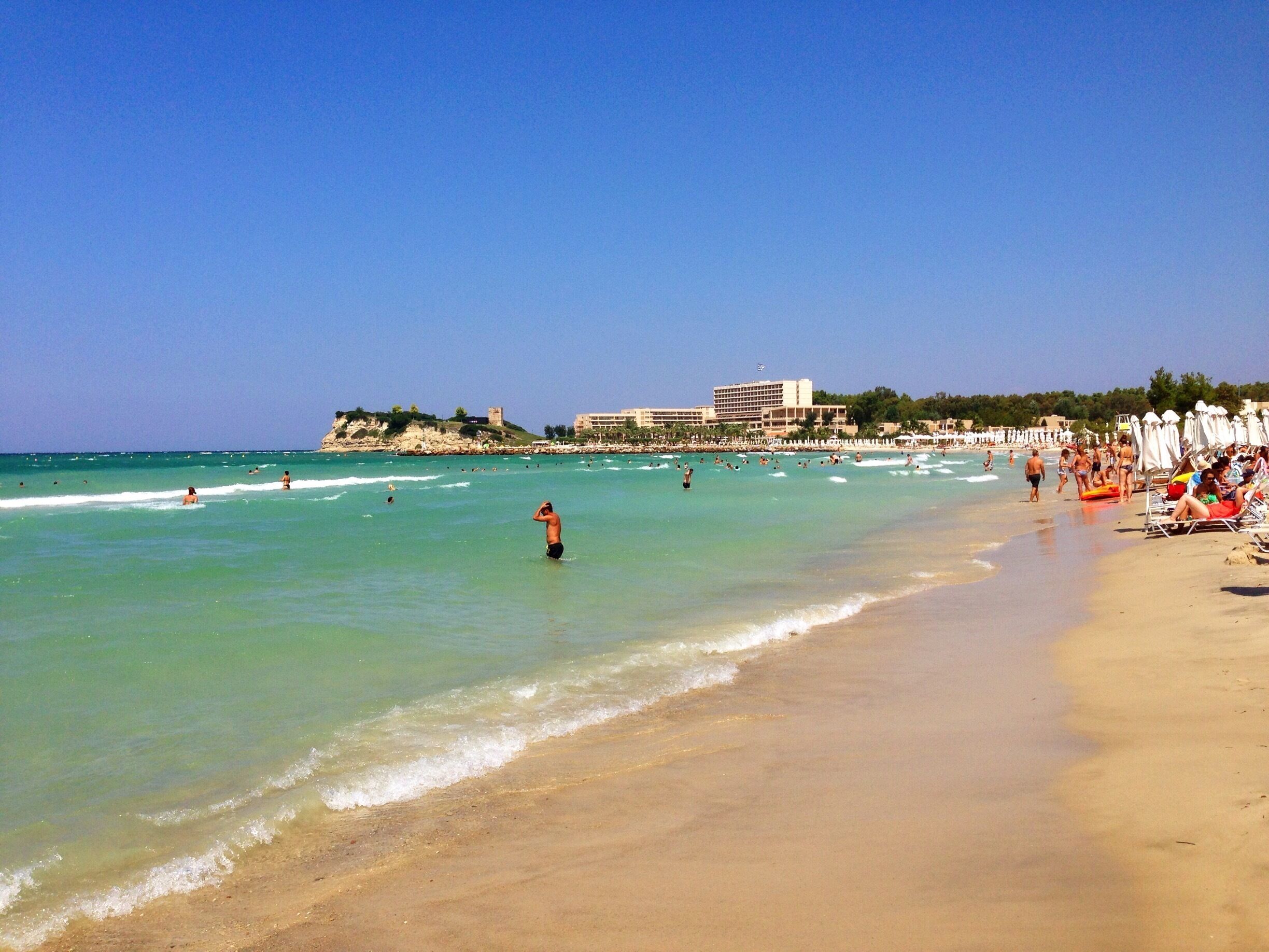 A beach with clear turquoise water, a sandy shore, and a castle-like structure in the background.