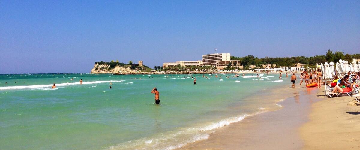 A beach with clear turquoise water, a sandy shore, and a castle-like structure in the background.