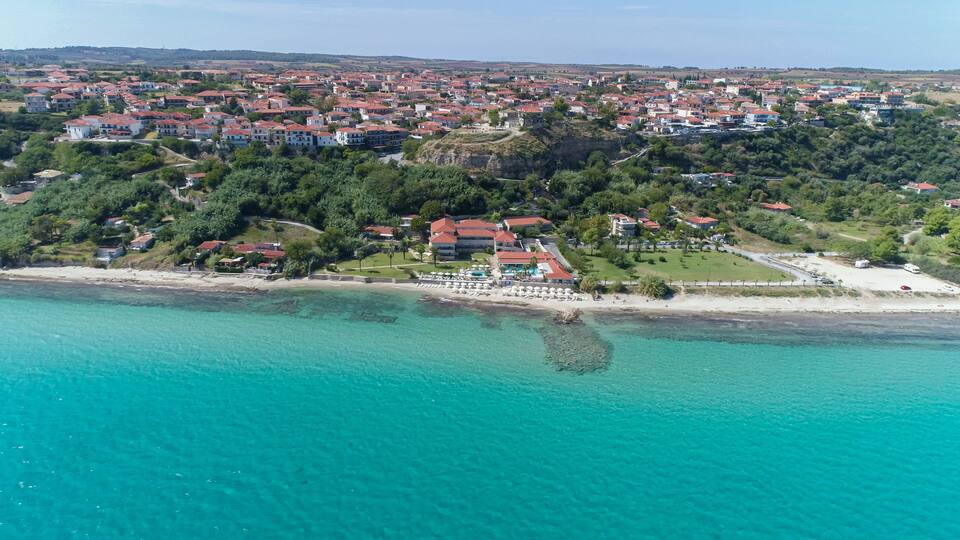 An aerial view of a coastal town with a beach, clear turquoise waters, and red-roofed buildings.