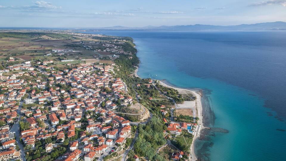 An aerial view of a coastal town with red-roofed houses, a beach, and a clear blue sea.