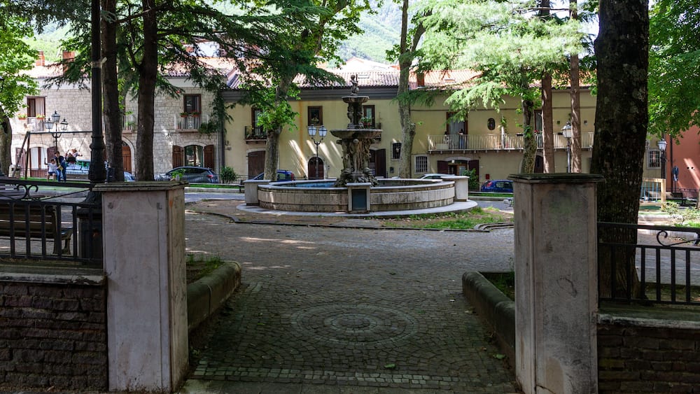 Une place pavée avec une fontaine, entourée d’arbres et de bâtiments historiques dotés de balcons.