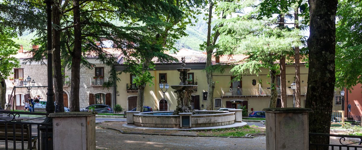 A cobblestone plaza with a fountain, surrounded by trees and historic buildings with balconies.