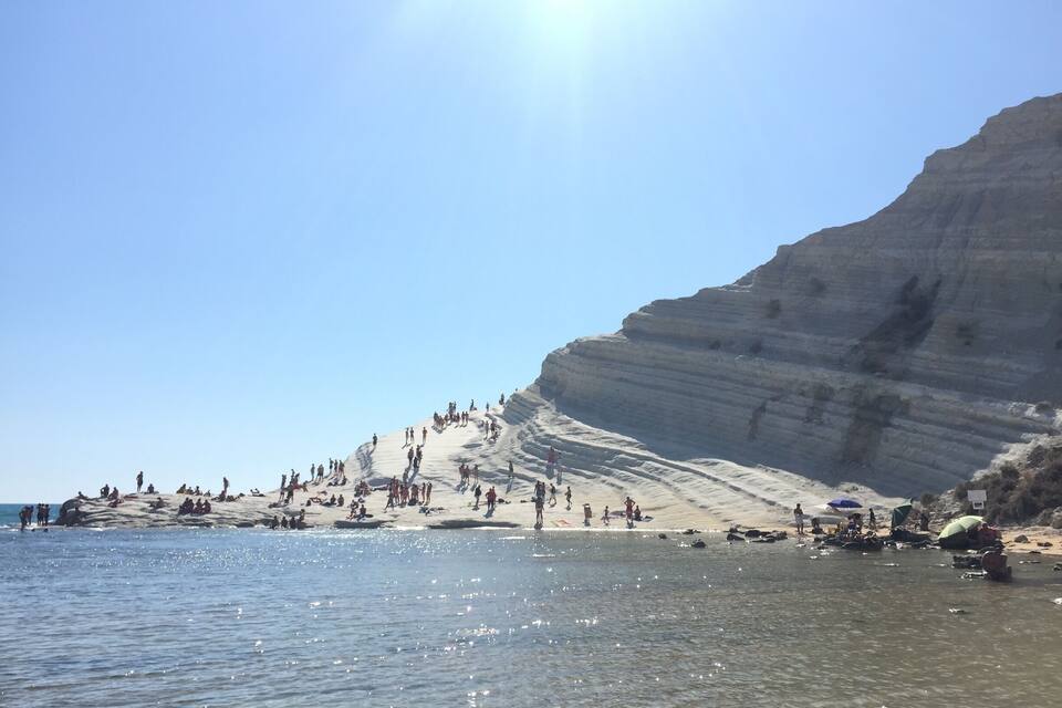 Una spiaggia con persone che prendono il sole e nuotano, un cielo sereno e un'alta scogliera sullo sfondo.