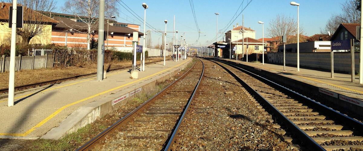 A railway station with tracks, overhead wires, and a clear blue sky.