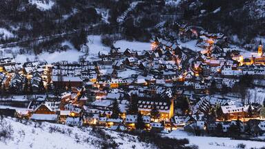 A snow-covered village at dusk with illuminated buildings and a prominent church steeple.