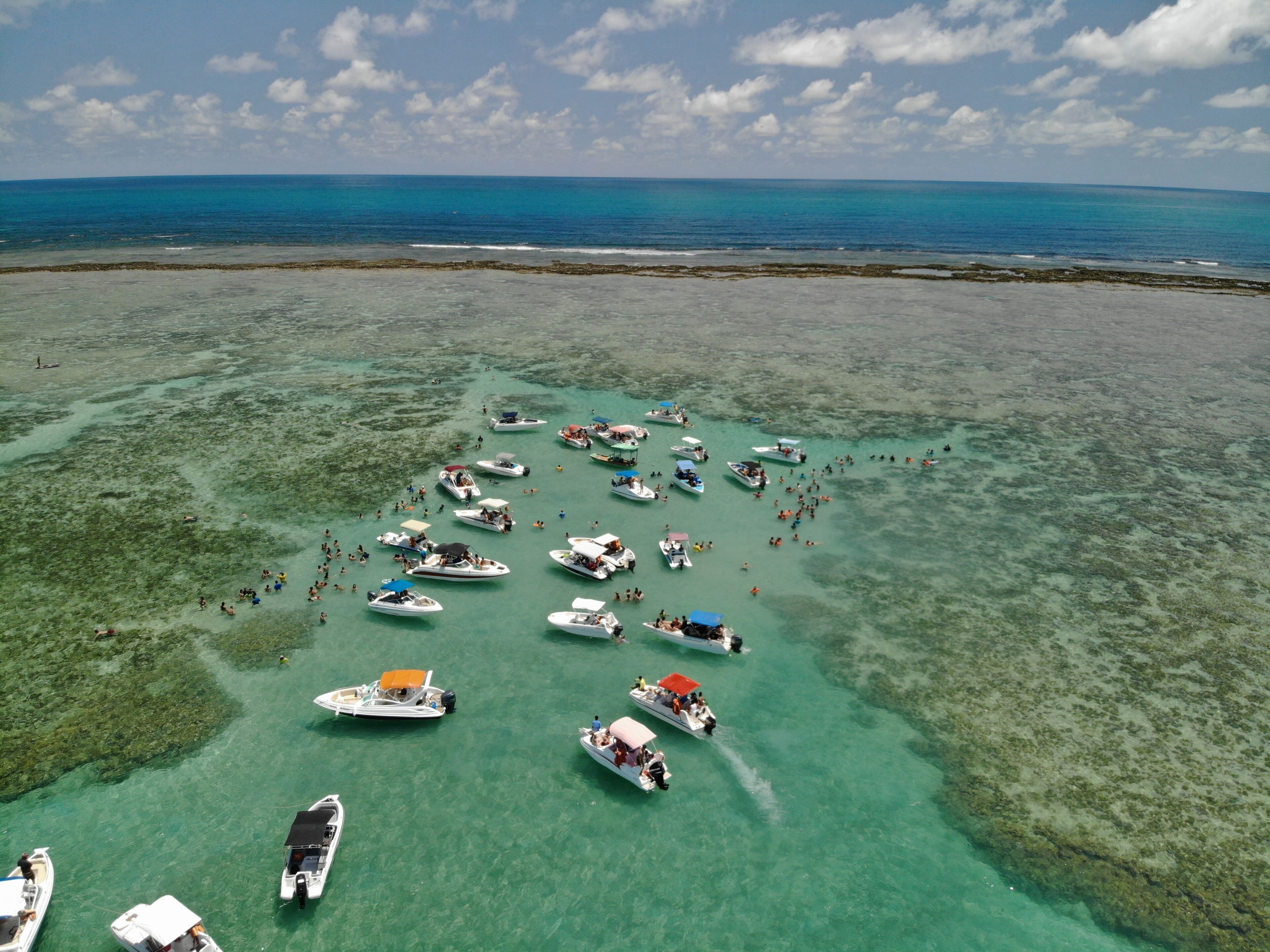 A tropical beach with numerous boats anchored in shallow waters, surrounded by clear turquoise sea and a distant shoreline.