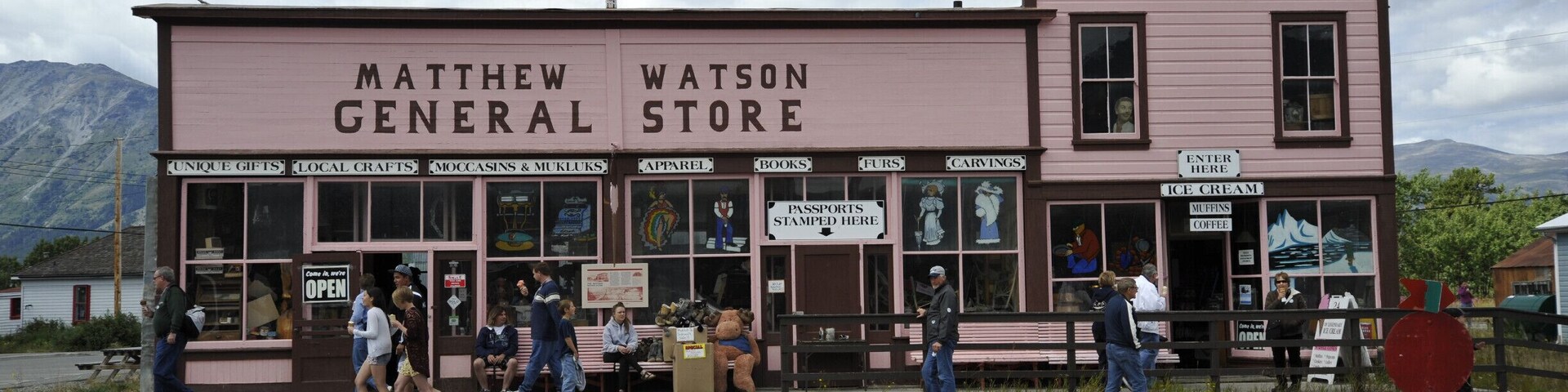 Un edificio rosa de dos pisos con la leyenda 'Matthew Watson General Store', con varios letreros que anuncian artículos como 'Regalos Únicos', 'Artesanías Locales' y 'Magdalenas y Café'.