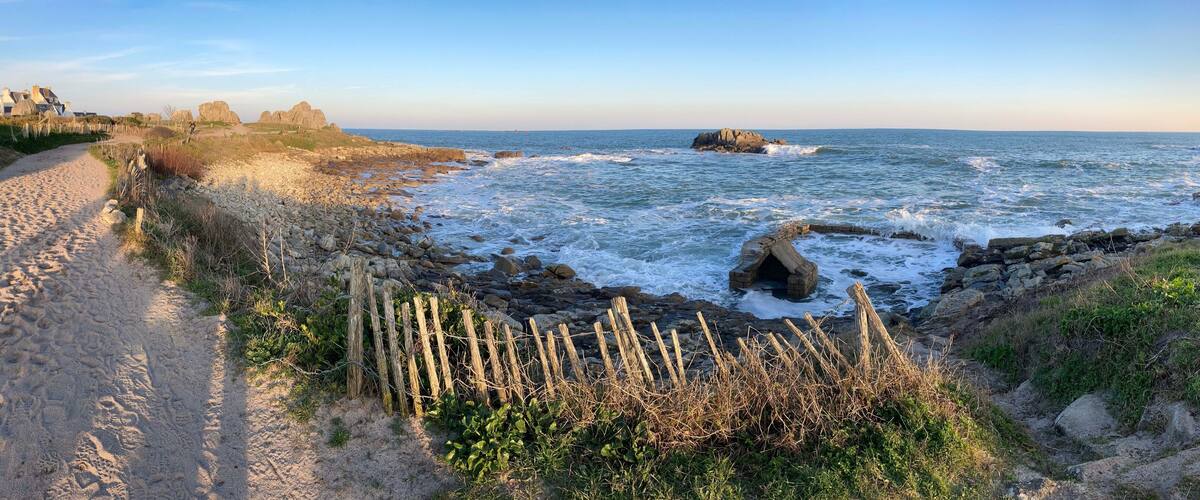 A coastal path with rocky terrain, a sandy beach, and a view of the ocean with waves crashing against rocks.