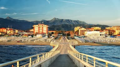 A coastal promenade with a bridge leading to a beach, surrounded by buildings and mountains.