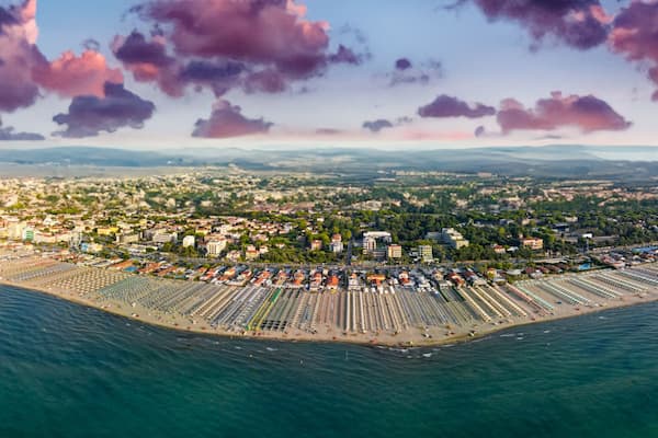 Una vista aerea di una città costiera con una zona urbana densamente popolata, una lunga spiaggia fiancheggiata da file di ombrelloni e lettini, e sullo sfondo montagne e un cielo al tramonto.