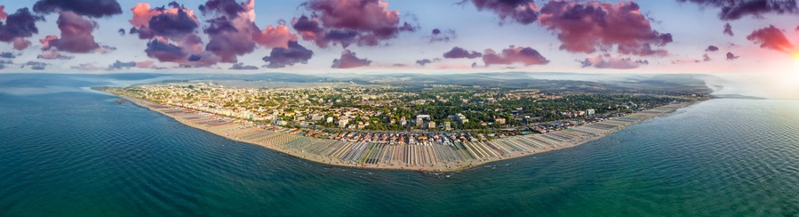 Una vista aerea di una città costiera con una zona urbana densamente popolata, una lunga spiaggia fiancheggiata da file di ombrelloni e lettini, e sullo sfondo montagne e un cielo al tramonto.