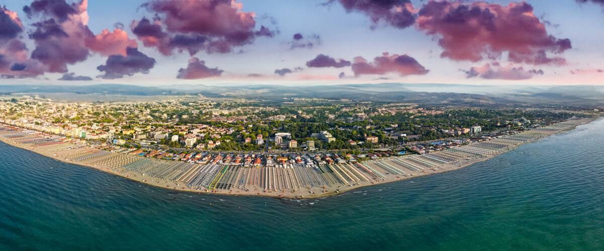 An aerial view of a coastal city with a densely populated urban area, a long beachfront lined with rows of beach umbrellas and sunbeds, and a backdrop of mountains and a sunset sky.